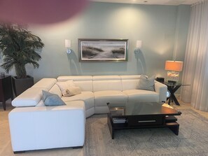 Ein modernes Wohnzimmer mit einem Ecksofa, einem Glastisch, einer Topfpflanze und einem gerahmten Landschaftsbild.