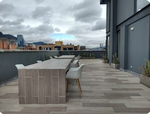 An outdoor terrace with a long table and chairs, surrounded by potted plants and a cityscape view. - Modern apartment with a view of central Chapinero (Bogotá)
