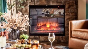 A living room with a fireplace, a television mounted on the wall, a brown leather sofa, a wooden coffee table with snacks and drinks, and a vase with dried flowers. - Private Pool • Smoky Mountain Views • Game Room (Gatlinburg)