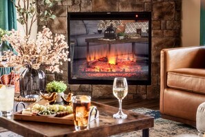 Ein Wohnzimmer mit Kamin, einem an der Wand befestigten Fernseher, einem braunen Ledersofa, einem Holztisch mit Snacks und Getränken sowie einer Vase mit Trockenblumen.