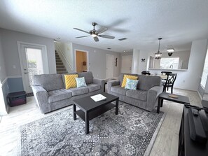 A living room with two grey sofas, a coffee table, a patterned rug, a ceiling fan, and a kitchen area with a dining table and chairs. - Golf Course Hideaway (Haines City)