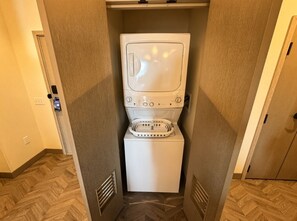 A compact laundry room with a front-loading washer and dryer, wooden flooring, and a door leading to another room. - Orlando | Couples Island Retreat | Pool + Beach (Golden Oak)