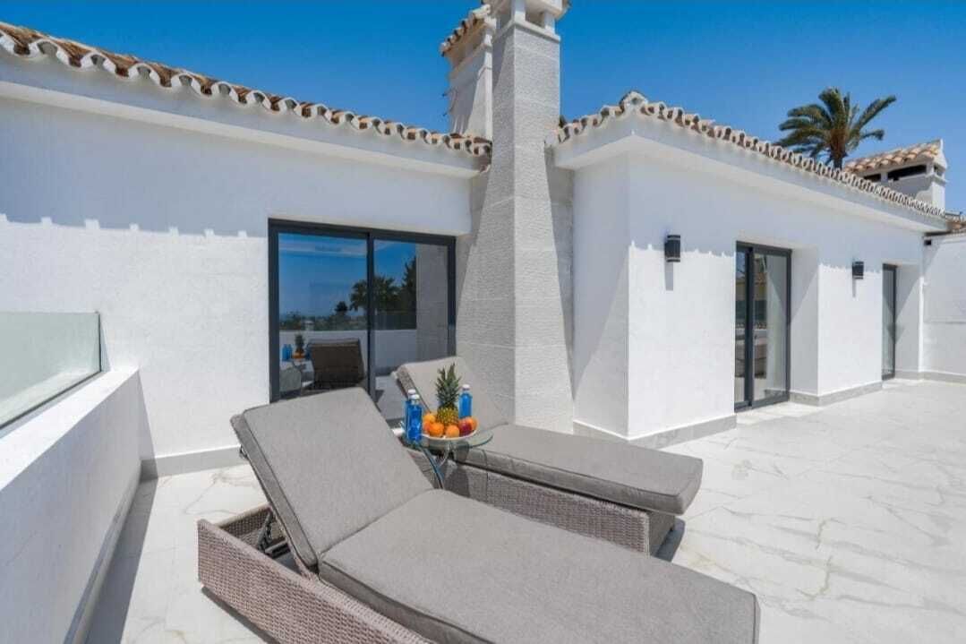 A rooftop terrace with lounge chairs, a table with fruit, and a building with a white facade and tiled roof.