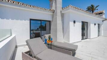 A rooftop terrace with lounge chairs, a table with fruit, and a building with a white facade and tiled roof.