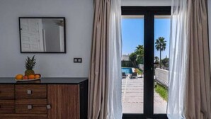 A room with a sliding glass door leading to an outdoor area, a wooden cabinet with drawers, a framed picture on the wall, and a fruit bowl on the cabinet. - Calle las camelias (Marbella)