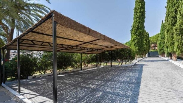 A covered outdoor area with a paved walkway, surrounded by trees and a clear sky. - Calle las camelias (Marbella)