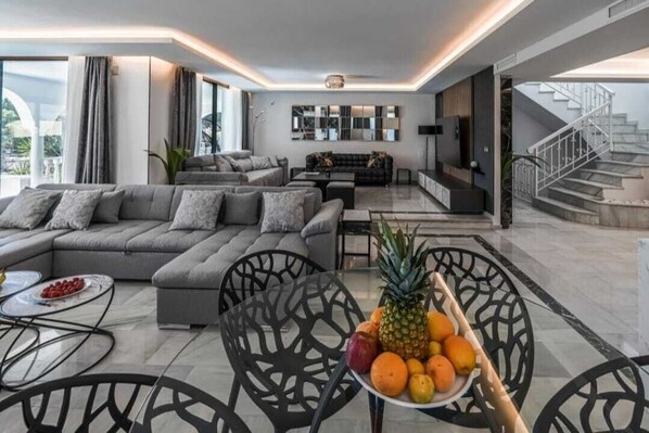 A modern living room with a sectional sofa, a dining table with chairs, and a fruit bowl.