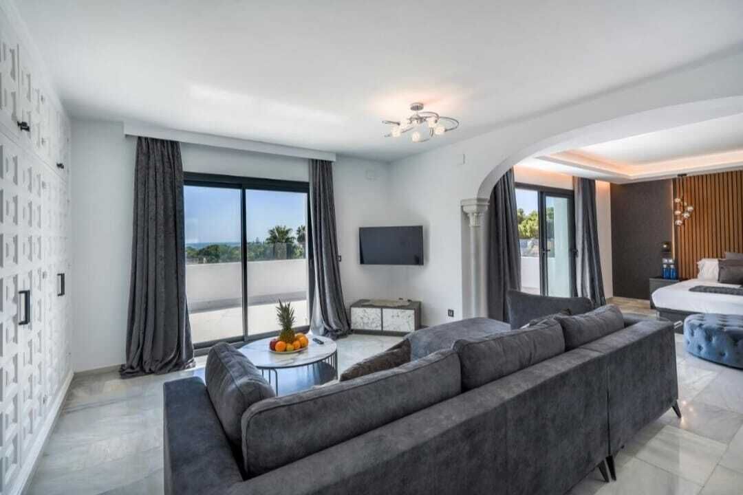 Een moderne woonkamer met een grijze bank, een klein rond tafeltje met fruit, en een flatscreen televisie aan de muur.