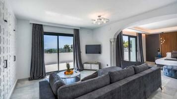 Een moderne woonkamer met een grijze bank, een klein rond tafeltje met fruit, en een flatscreen televisie aan de muur.