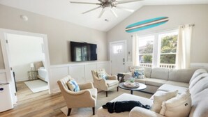 A living room with a ceiling fan, a sofa, armchairs, a coffee table, and a television. - Charming Oak Island Retreat — Minutes from the Beach with Private Hot Tub (Oak Island)
