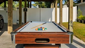 A pool table with a wooden frame and a cue stick, surrounded by a white fence and greenery. - Charming Oak Island Retreat — Minutes from the Beach with Private Hot Tub (Oak Island)