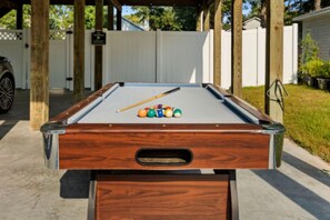 Ein Billardtisch mit Holzrahmen und Spielstange, umgeben von einem weißen Zaun und Grünpflanzen.