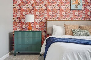 A bedroom with a floral wallpaper, a green dresser, a white bed with a blue blanket, and a framed picture on the wall.