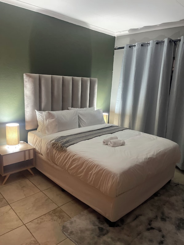 A neatly made bed with a headboard, a bedside lamp, and a fluffy rug on a tiled floor.