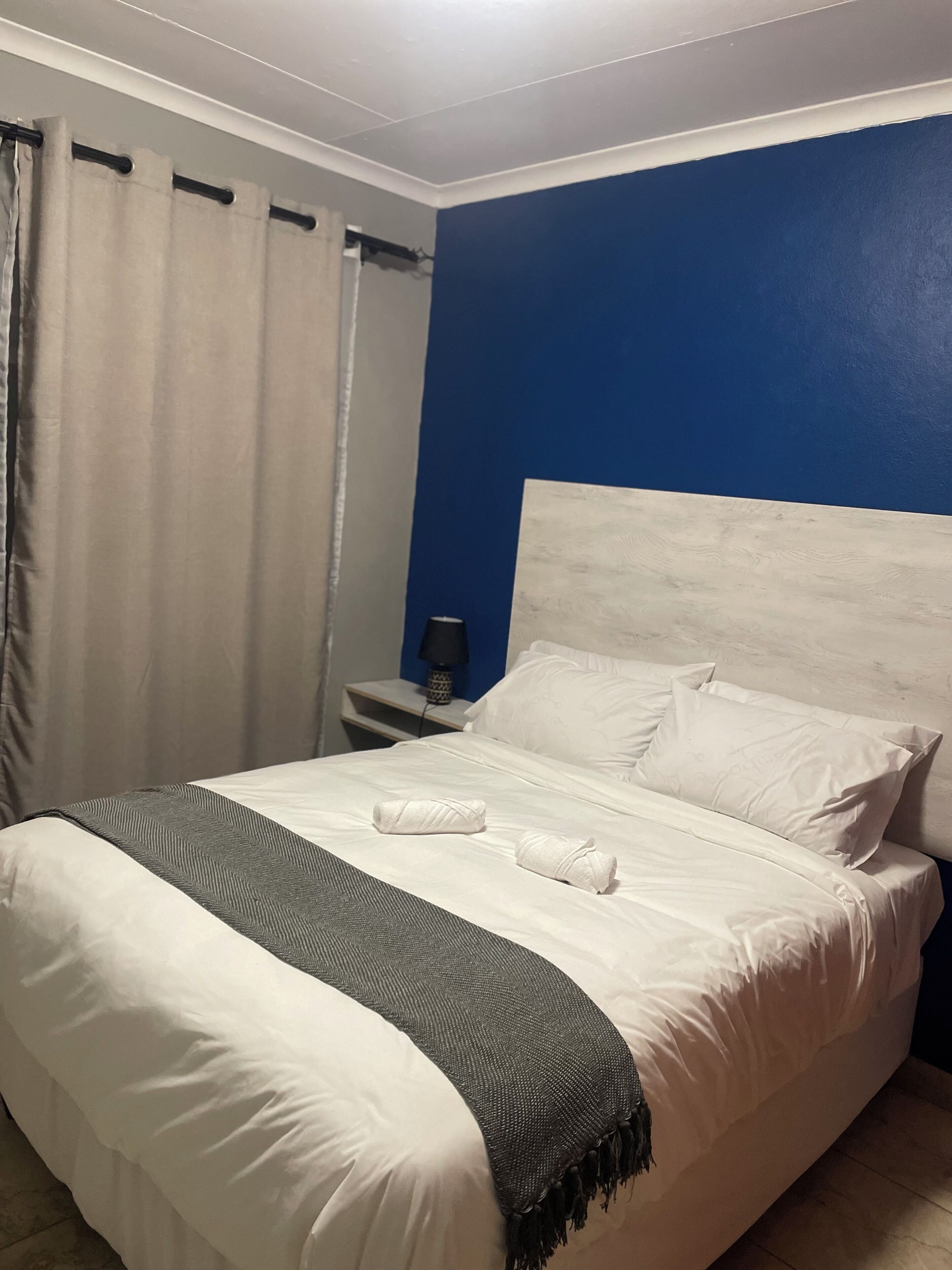 Quarto de hotel com cama, roupa de cama branca, parede de destaque azul escuro, mesa de cabeceira com abajur e cortina.