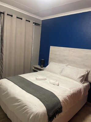 A hotel room with a bed, white bedding, a dark blue accent wall, a nightstand with a lamp, and a curtain.