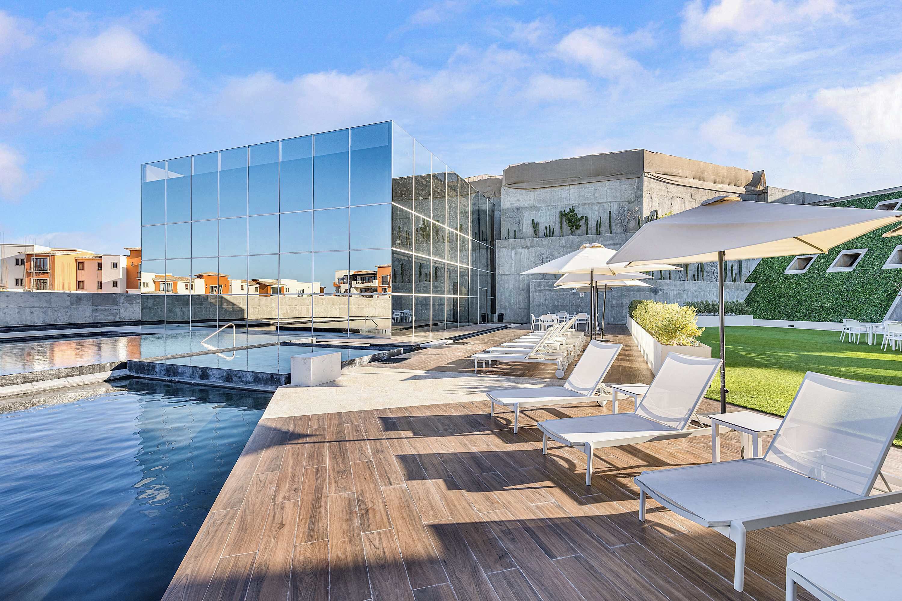 Un área de piscina en la azotea con sillones de descanso y sombrillas, un edificio moderno con fachada de vidrio reflectante y vistas de la ciudad.