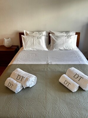 A neatly made bed with white linens and pillows, a bedside lamp, and two rolled towels with 'DT' on them.