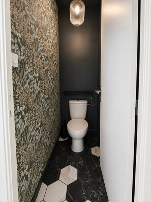 Ein schmales Badezimmer mit Toilette, einer Wandleuchte und einem gemusterten Wandpapier.