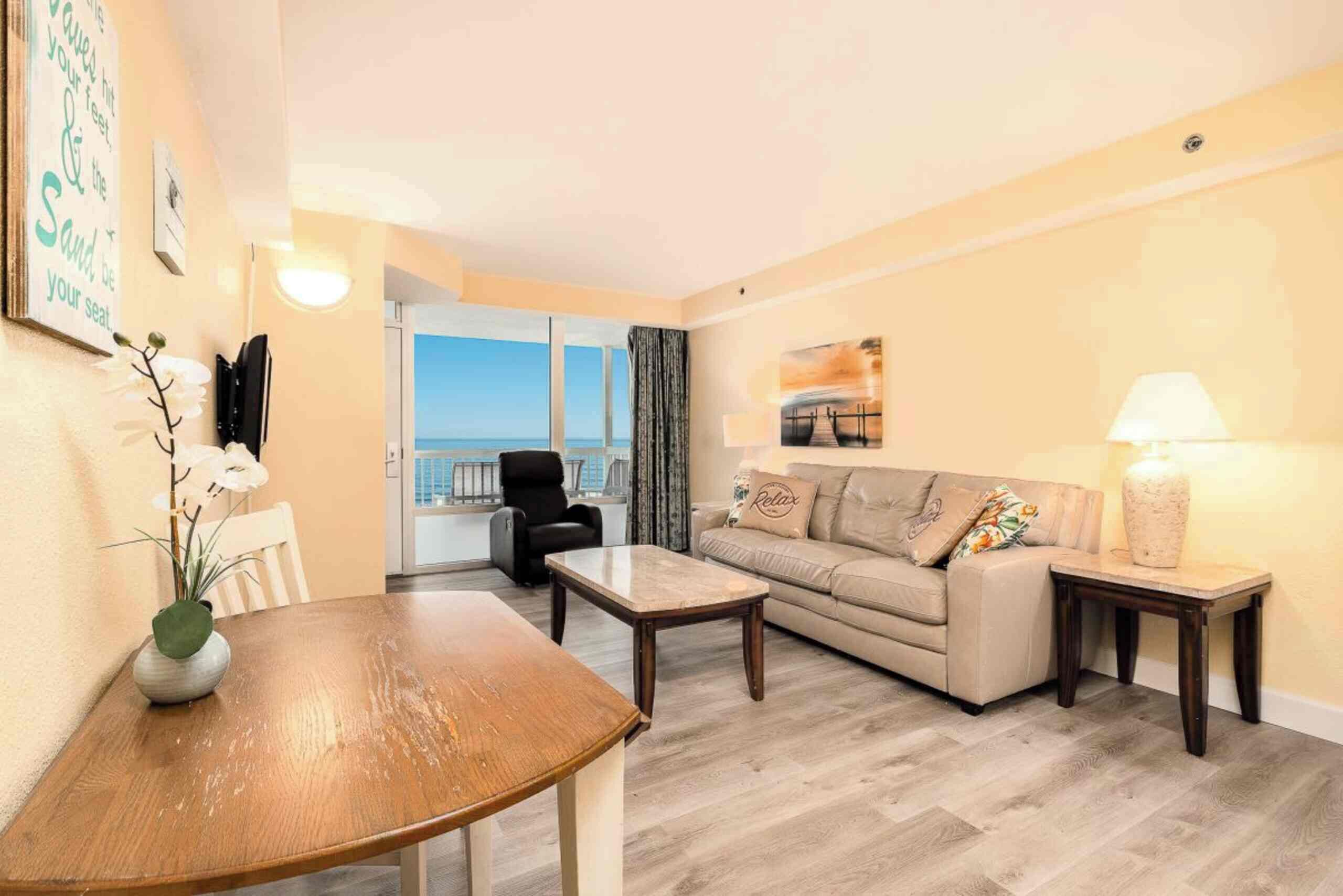 A spacious living room with a sofa, a coffee table, and a dining table. There is a view of the sea through large windows.