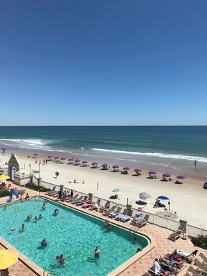 Ett poolområde med kristallklart blått vatten, omgivet av solstolar och parasoller, med utsikt över en sandstrand och havet.