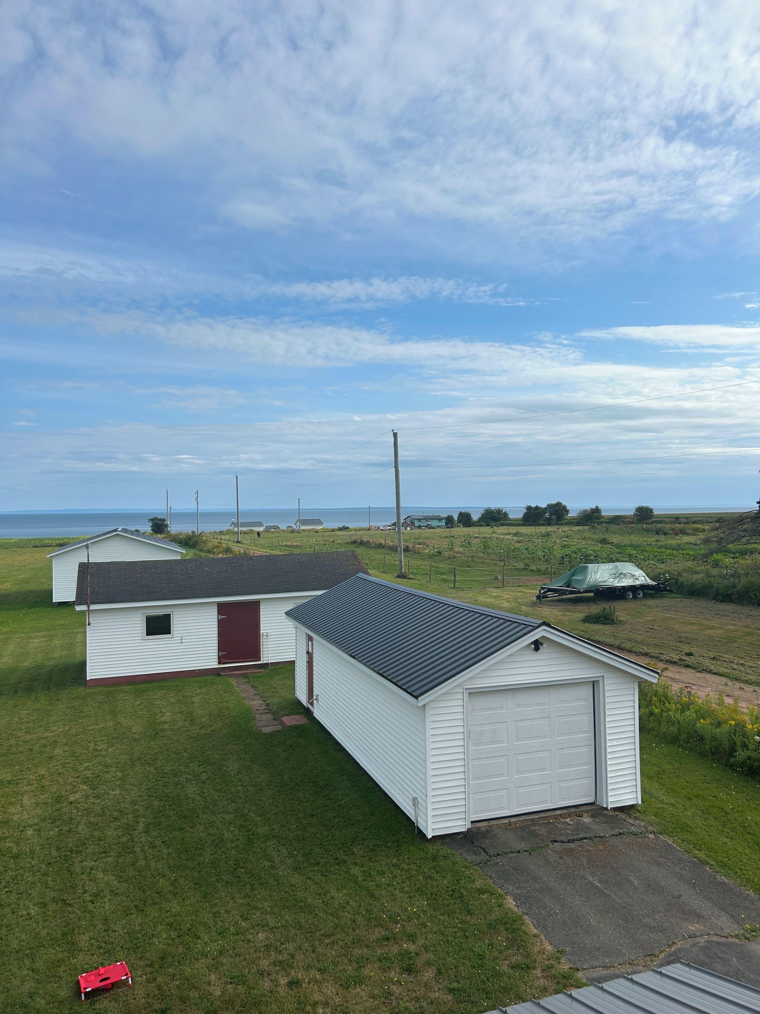 A white building with a red garage door, a small shed, and a trailer with a green tarp.