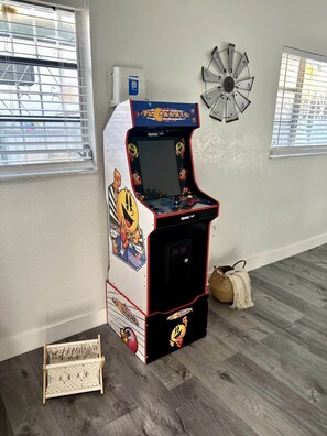 A Pac-Man arcade machine in a room with wooden flooring, a decorative wall clock, and a window with blinds. - Lovely Home in Little Havana- Near Loan Depot Park (Miami)