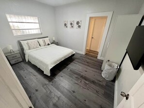 A bedroom with a bed, a nightstand, a window, a door, and wall art. - Lovely Home in Little Havana- Near Loan Depot Park (Miami)