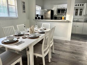 Een eetgedeelte met een witte tafel gedekt voor vier personen, een keuken met moderne apparatuur, en een uitzicht op het landschap door een raam.