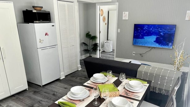 Una zona de dinar amb taula preparada per a quatre, microones, nevera i televisió penjada a la paret.