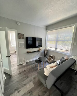 Een moderne woonkamer met een grijze bank, een flatscreen televisie, een kleine tafel en een raam met jaloezieën.