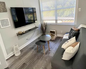 Ein modernes Wohnzimmer mit Fernseher, rundem Couchtisch, grauem Sofa und Kissen mit Muster sowie einem Fenster mit Jalousien.