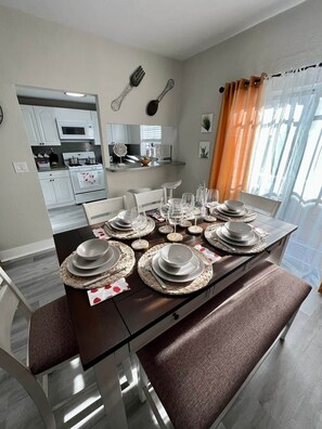 Un comedor con una mesa dispuesta para cuatro, una cocina al fondo y una ventana con cortinas.