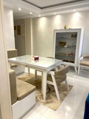 Ein modernes Wohnzimmer mit einem Esstisch mit Glasplatte, beigefarbenen Sofas, einem gemusterten Teppich und einer weißen Kommode mit Glastüren.