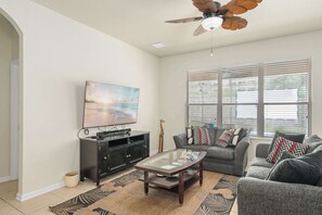 A living room with a television, a ceiling fan, a sofa, a coffee table, and a window with blinds. - Themed House with Heated Private Pool & Game Room (Kissimmee)