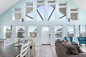 A spacious room with a high ceiling, large windows, a dining area, and a seating area with a sofa and chairs. - The Luxe Coast House -  Walk to the Beach (Bolivar Peninsula)