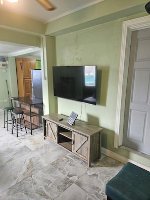 Ein Wohnzimmer mit Flachbildfernseher an einem Holzkasten, eine Essecke mit Tisch und Stühlen sowie eine Küchenzeile mit Kühlschrank.