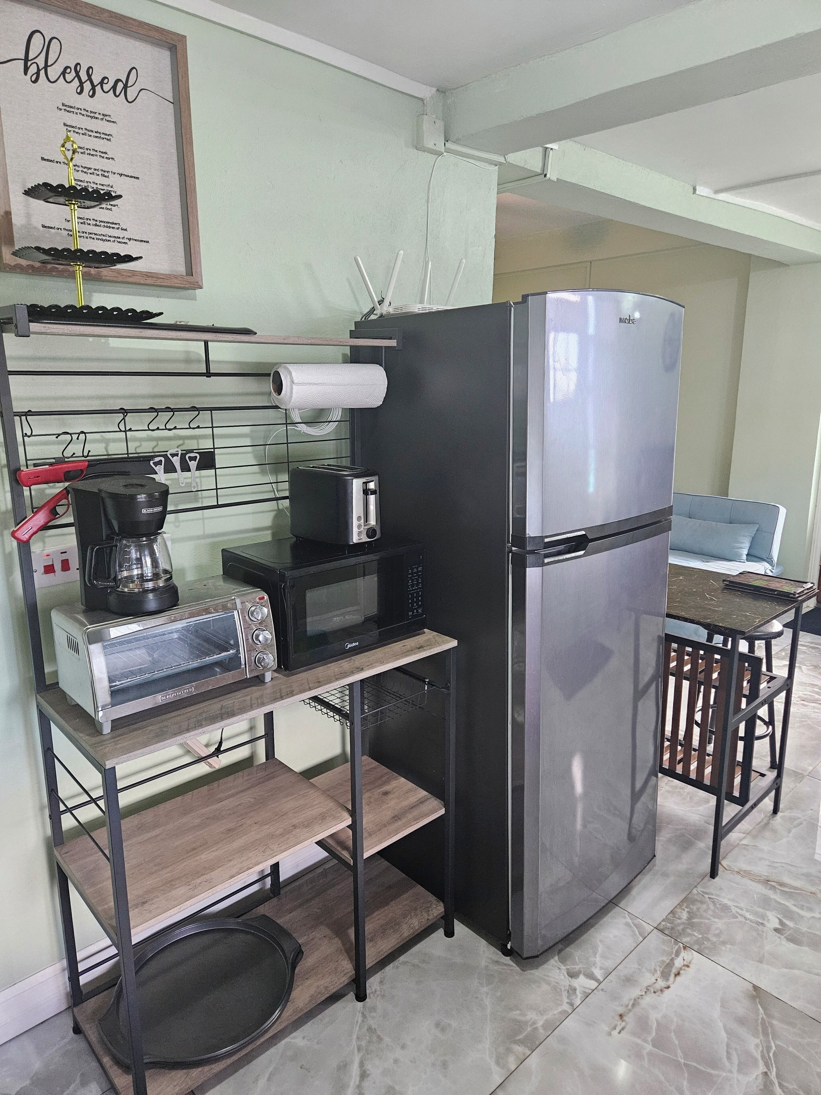 A compact kitchen with a stainless steel refrigerator, a microwave, a toaster, and a coffee maker on a metal rack.
