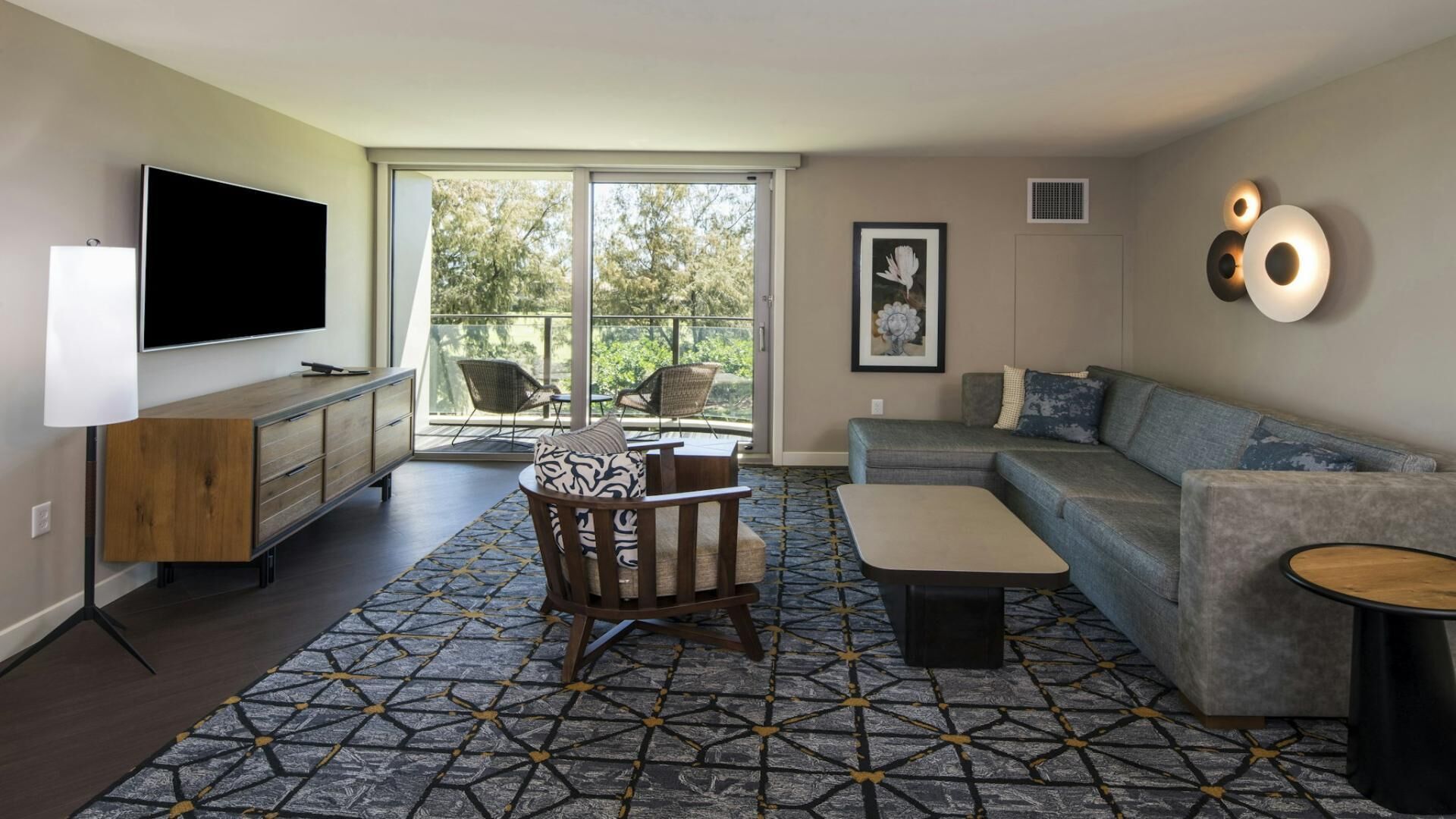 Un salon moderne doté d’un téléviseur à écran plat, d’un canapé, d’un tapis à motifs et d’un balcon avec des chaises.