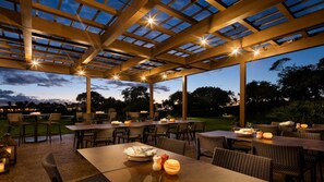 Ein Außenbereich mit Terrasse, Tischen, Stühlen, einer Pergola mit Beleuchtung und Blick auf die Landschaft.