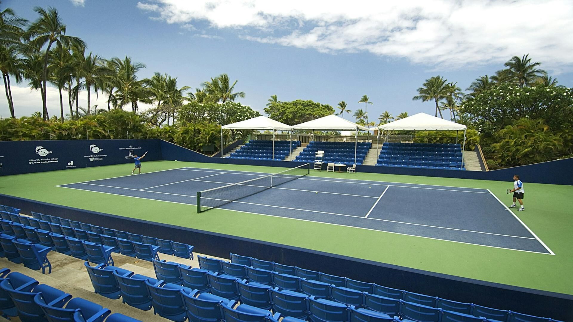 Una cancha de tenis con asientos azules, toldos blancos y palmeras al fondo.