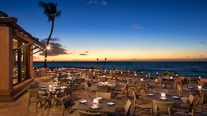 Un espace extérieur aménagé pour les repas, avec des tables et des chaises, offrant une vue sur l’océan au coucher du soleil.