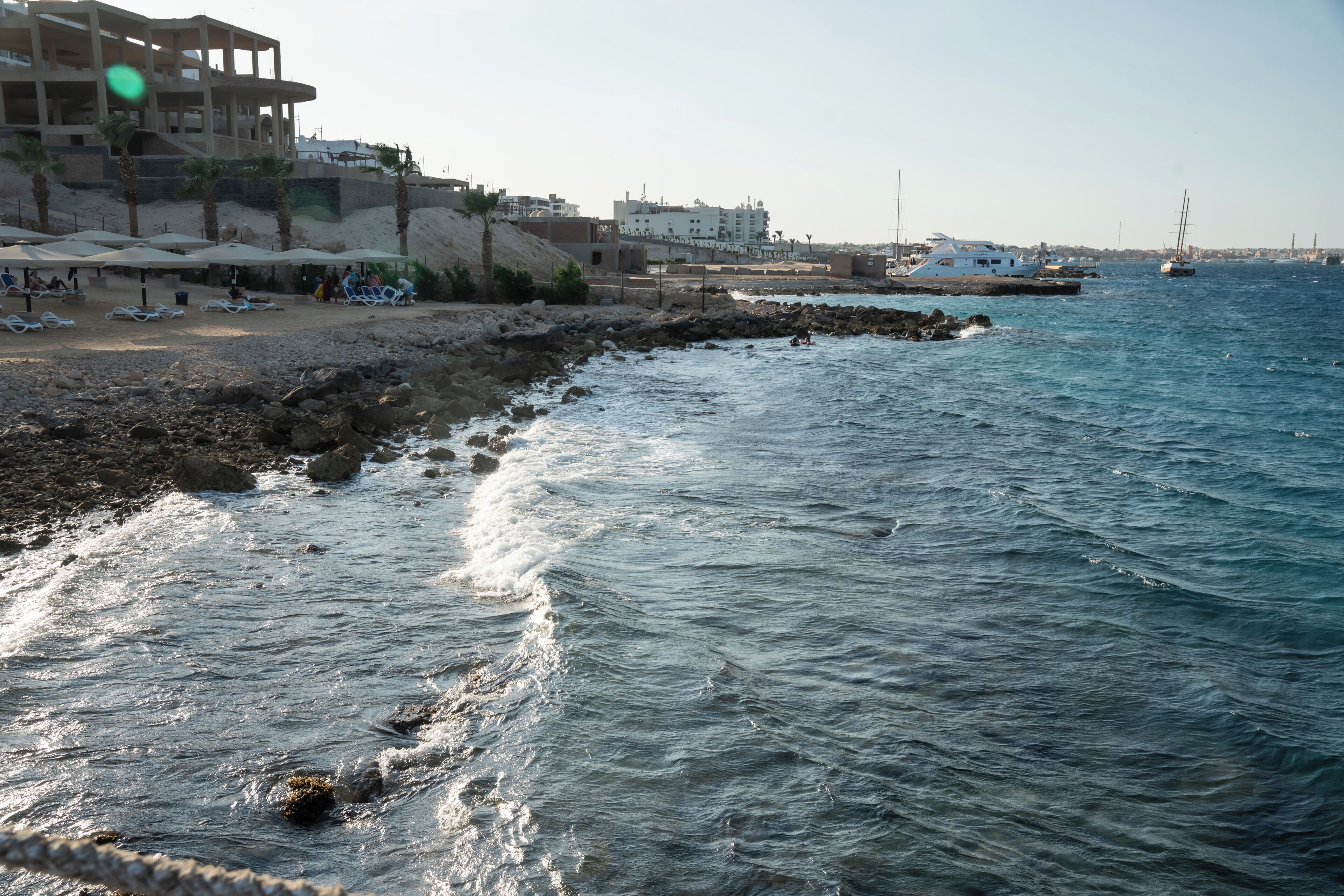 A coastal area with a rocky shoreline, a sandy beach with umbrellas, and a view of the sea with boats.