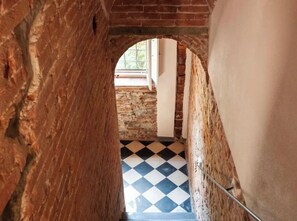 Een smalle doorgang met een zwart-wit schaakbordvloer die leidt naar een kamer met een bakstenen muur en een raam.
