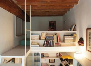 Ein kleines Zimmer mit Schlafplatz in der Schräge, einem Bücherregal und einem kleinen Tisch mit Lampe.