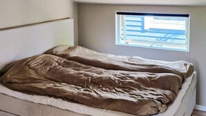 Una cama individual con colchón color crema y manta marrón en una habitación con ventana pequeña.