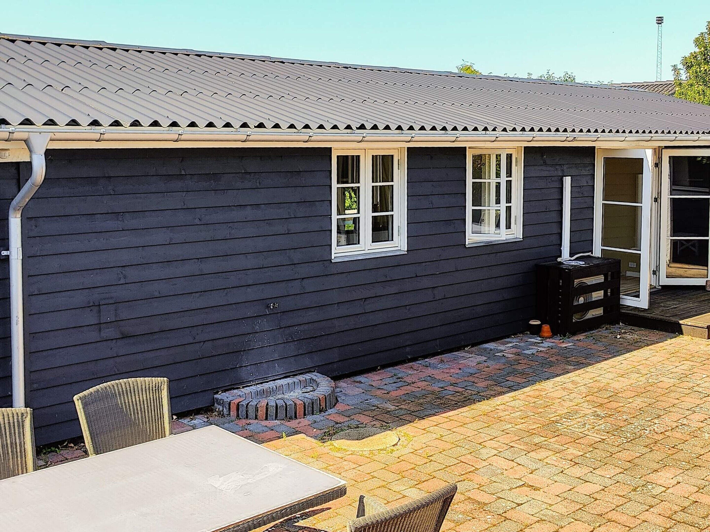 A dark wooden building with a metal roof, a covered patio area with a table and chairs, and a door leading to an interior space.