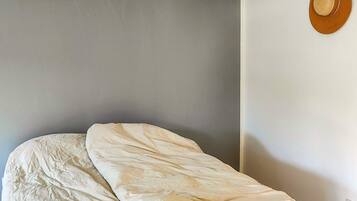 Una cama con edredón blanco en una habitación con pared gris lisa y un sombrero colgado en la pared.