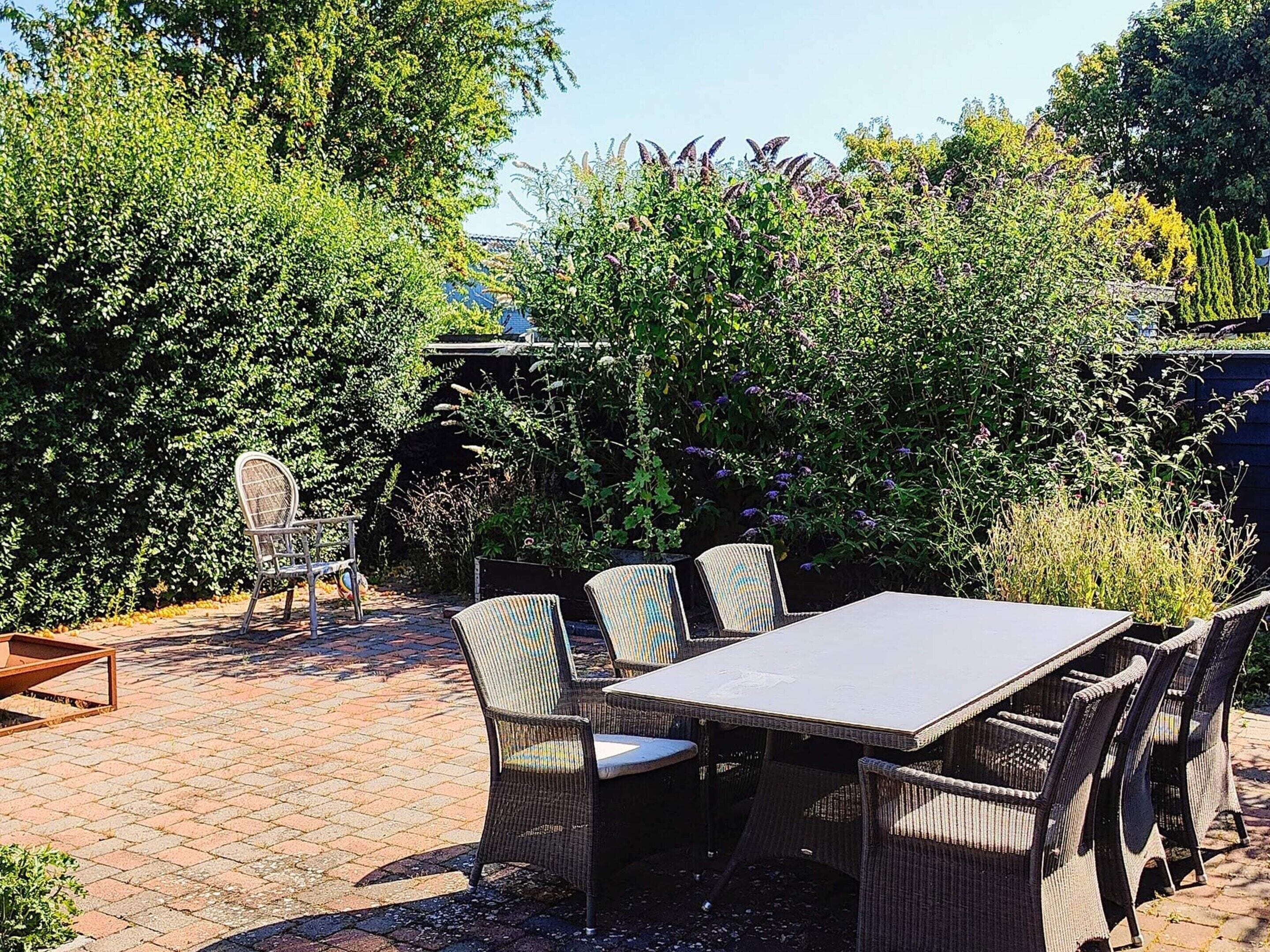 An outdoor patio with a table and chairs, surrounded by dense foliage and a brick pathway.