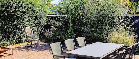 Eine Terrass mit Tisch und Stühlen, umgeben von dichter Bepflanzung und einem Ziegelweg.